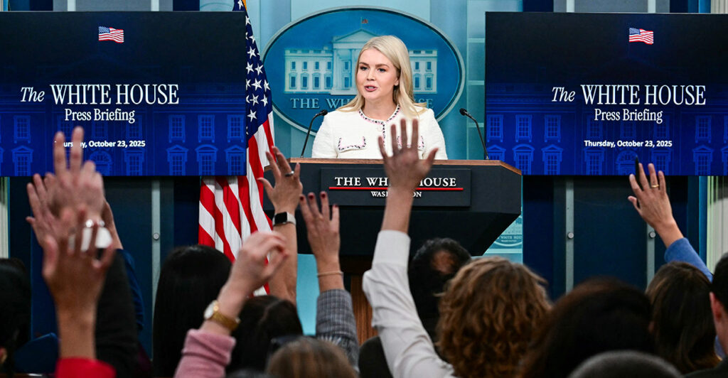 Karoline Leavitt in white outfit address reporters, who have their hands raised.