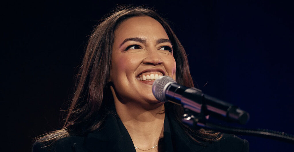 Alexandria Ocasio-Cortez smiles as she stands before a microphone.