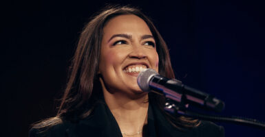 Alexandria Ocasio-Cortez smiles as she stands before a microphone.