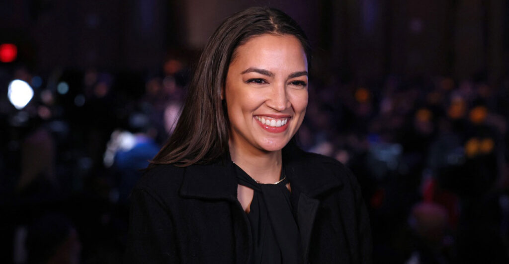 Alexandria Ocasio-Cortez stands at an event and smiles while she celebrates.