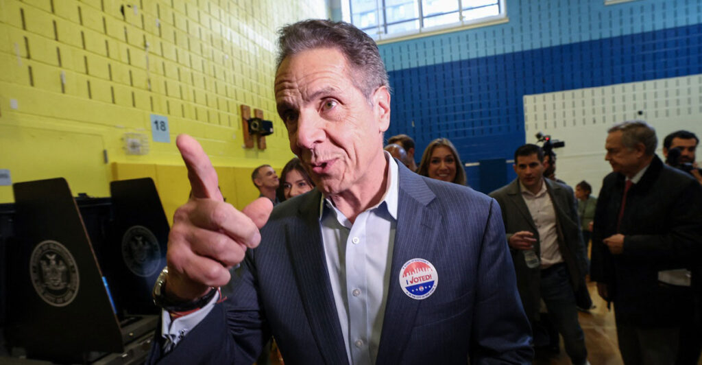 Andrew Cuomo stands in a room and points with his right hand while talking.