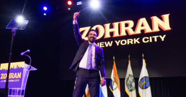 Zohran Mamdani gives a thumbs up sign with his right hand as he walks across a stage with a smile on his face.