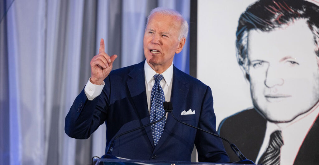 Joe Biden stands at a podium while talking and holds his right hand up in the air.