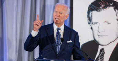 Joe Biden stands at a podium while talking and holds his right hand up in the air.