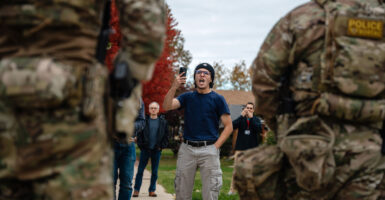 Two ICE agents with back to camera are confronted by an activist in black shirt and grey pants, holding a cellphone.