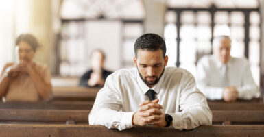 Male in white shirt and tie sits in wooden pew praying.