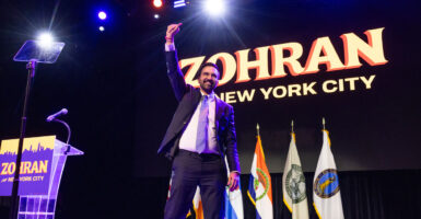 Zohran Mamdani raises a fist on a stage backed with a sign reading "ZOHRAN New York City."