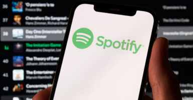 Spotify logo on a smartphone held by hand, with list of artists in the background.