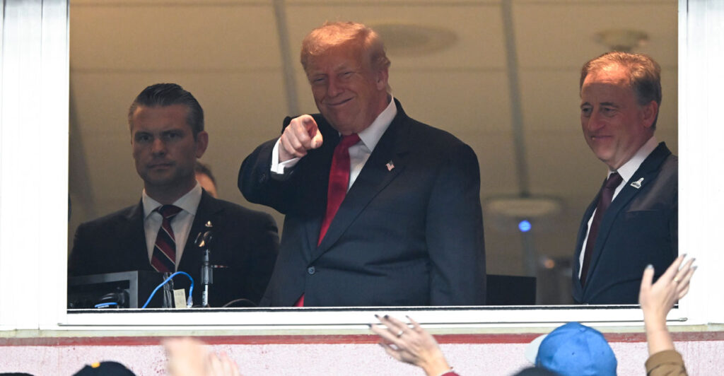 President Trump in the Commanders owner's box smiles and points at crowd.