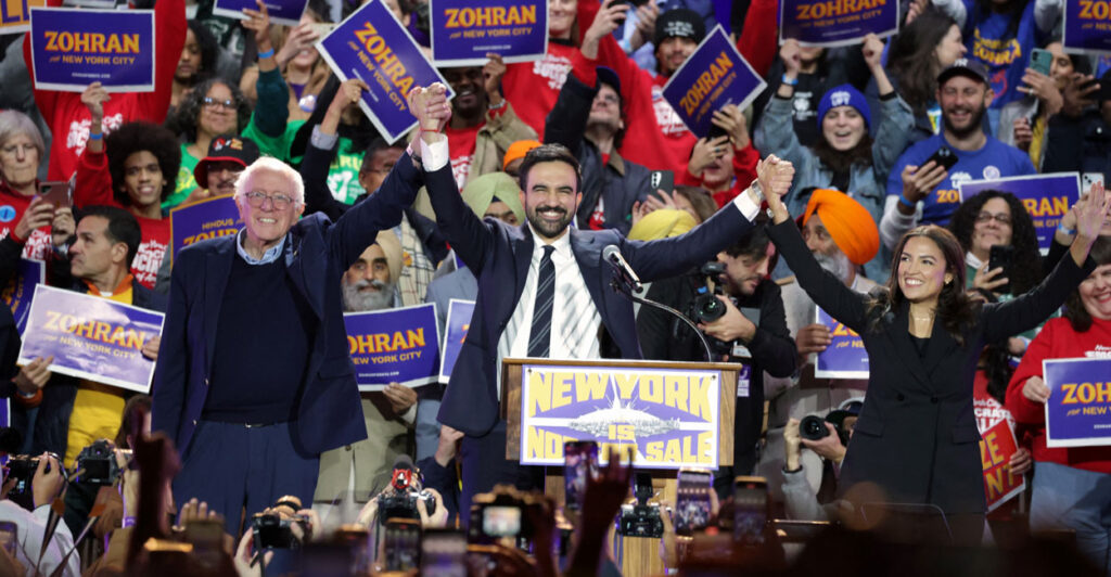 Zohran Mamdani raises the arms of Bernie Sanders and Alexandria Ocasio-Cortez, with crowd behind them.
