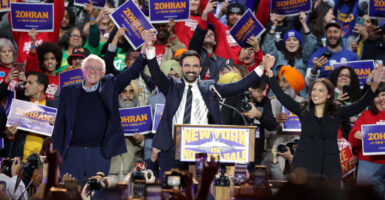 Zohran Mamdani raises the arms of Bernie Sanders and Alexandria Ocasio-Cortez, with crowd behind them.