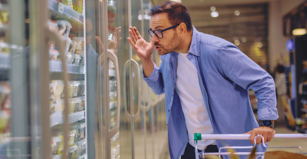 Man in blue shirt, white T-shirt, and glasses appears to be yelling at items in grocery store fridge.