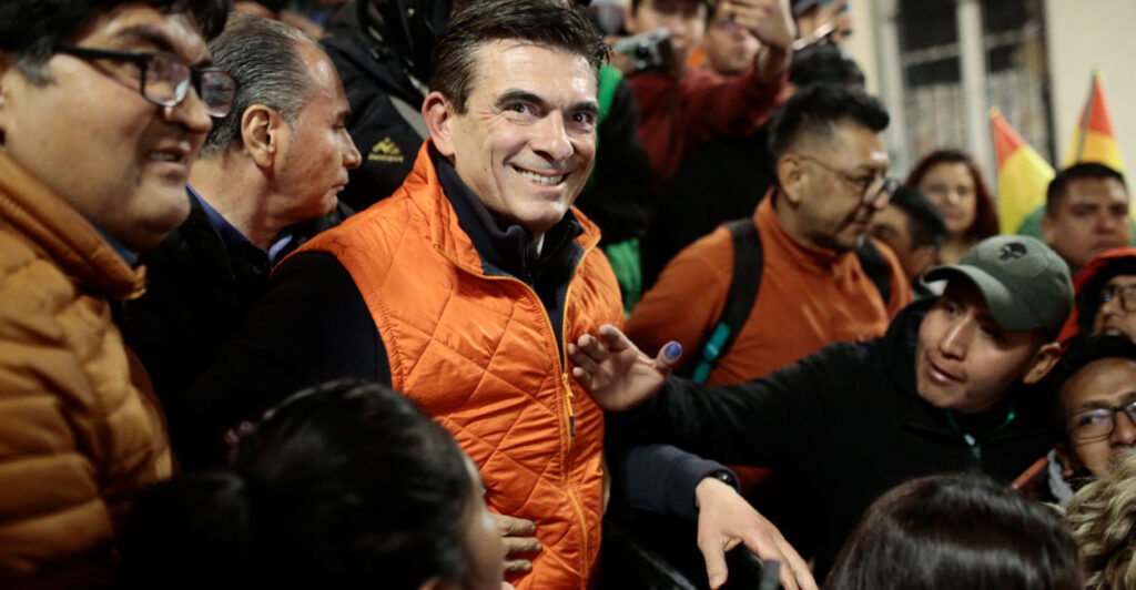 Rodrigo Paz in an orange down vest smiles as he is surrounded by supporters.