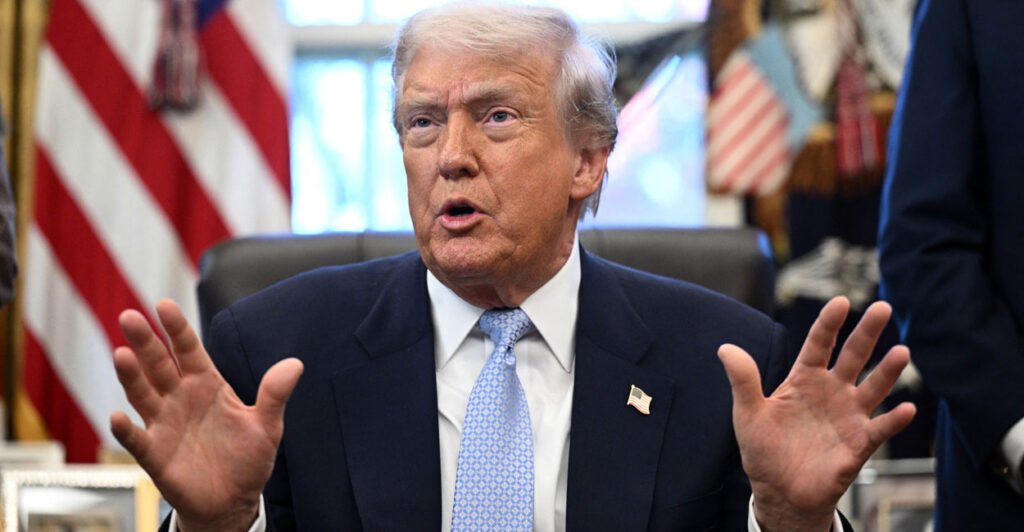 Donald Trump at his desk in the Oval Office, his hands in front of him.