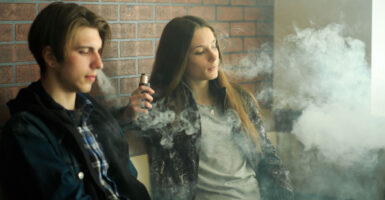 A young male and female sit along a brick wall vaping.