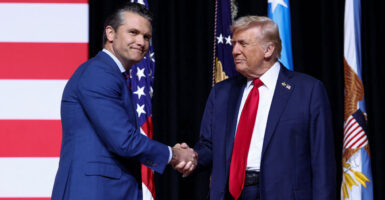 Secretary of War Pete Hegseth shakes hands with President Trump on a stage in front of American flags.