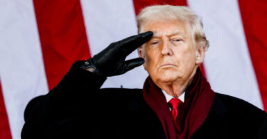 Close-up of President Trump, in a black winter coat, saluting, backed by large American flag.