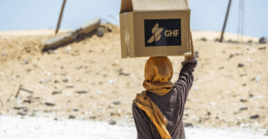 Woman, her head wrapped, balances and carries on her head a box of aid from Gaza Humanitarian Foundation.