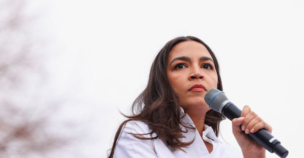 Alexandria Ocasio-Cortez in a white blouse behind a microphone, with a pale, outdoor background.