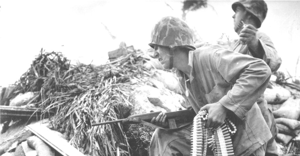 During the Battle of Tarawa, a United States Marine, an ammunition belt in one hand and a carbine rifle in the other, as he crouches beside a soldier who rears back to throw a grenade, Tarawa, Kiribati, 1943.
