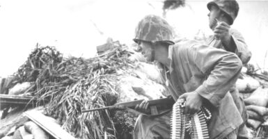 During the Battle of Tarawa, a United States Marine, an ammunition belt in one hand and a carbine rifle in the other, as he crouches beside a soldier who rears back to throw a grenade, Tarawa, Kiribati, 1943.