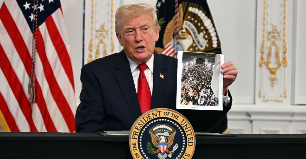 President Trump speaks to the U.S. military, with a backdrop of a white wall with elaborate gold trim. He's holding up a photo of a cargo plane full of Afghan refugees.