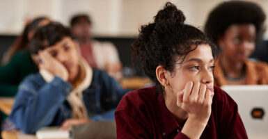 Bored teenage female looks out classroom window while male classmate behind her is fast asleep.