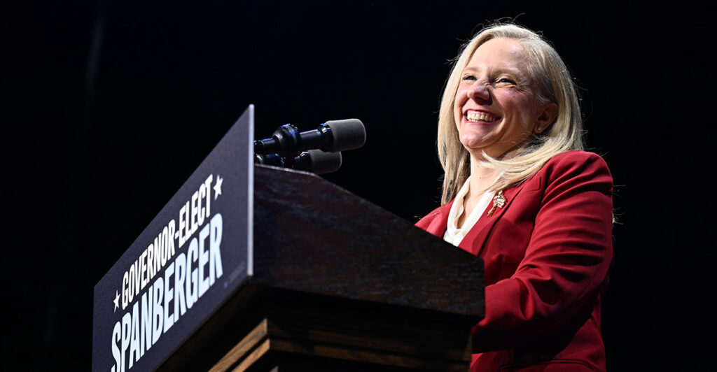 Abigail Spanberger in red smiles behind a podium. Spanberger is likely to reverse at least 6 Youngkin policies, and likely more.