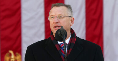 Veterans Affairs Secretary Doug Collins in a winter overcoat speaks in front of a large American flag.