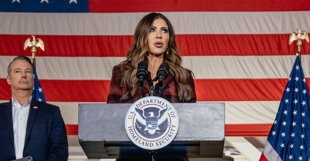 Kristi Noem at a podium in front of a large American flag.