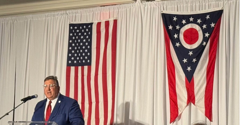 Ohio GOP Chairman Alex Triantafilou stands at a podium with the American and Ohio flags behind him.