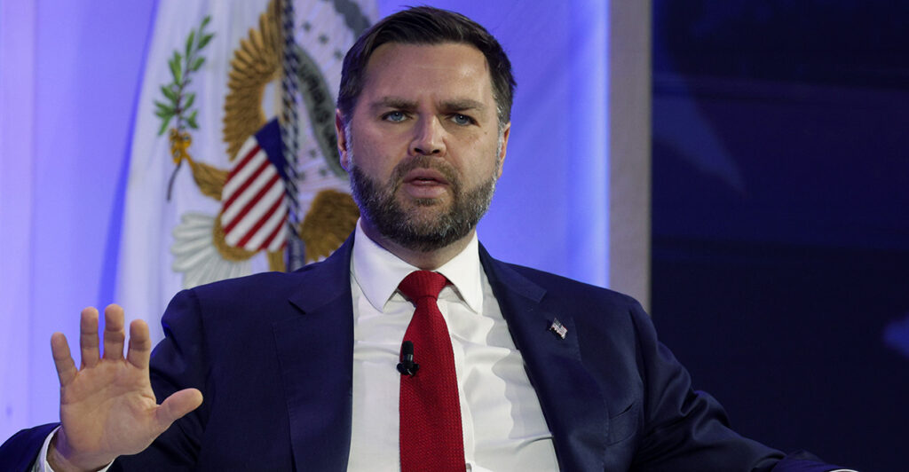 JD Vance seated, in a blue suit with red tie, with his right hand up.