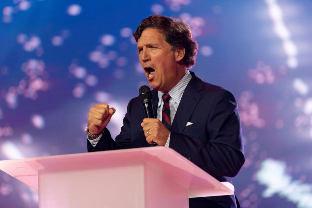 Tucker Carlson with fists clenched speaks at pink podium.