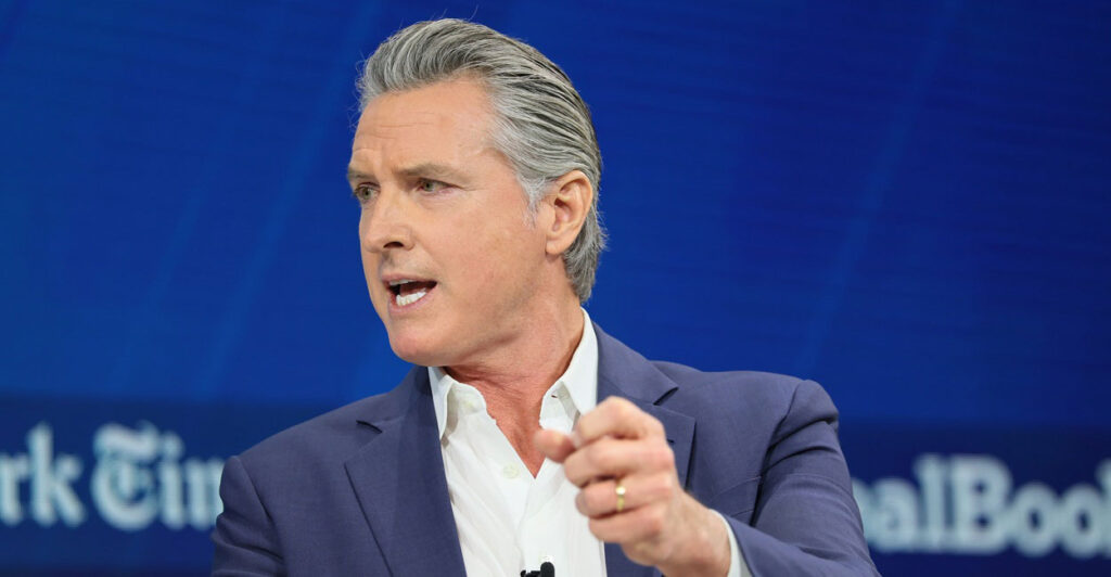 Gavin Newsom sits in a chair and looks to his right while gesturing with his left hand.