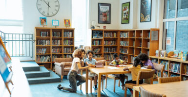 A woman kneels at a table where a young child is reading a book in a library.