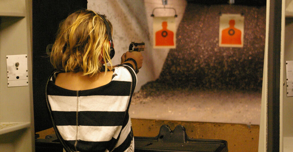 Blonde woman in black and white shirt fires handgun at an indoor shooting range.