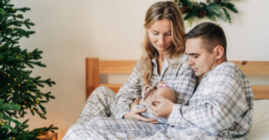Parents in grey and white pajamas sit on their bed, holding a newborn. A Christmas tree is on the left side of the shot.