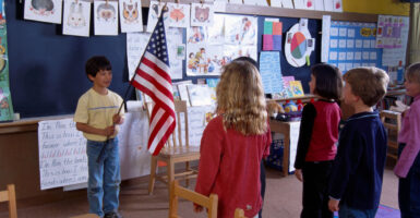 Little children stand in their classroom in front of a boy holding an American flag.