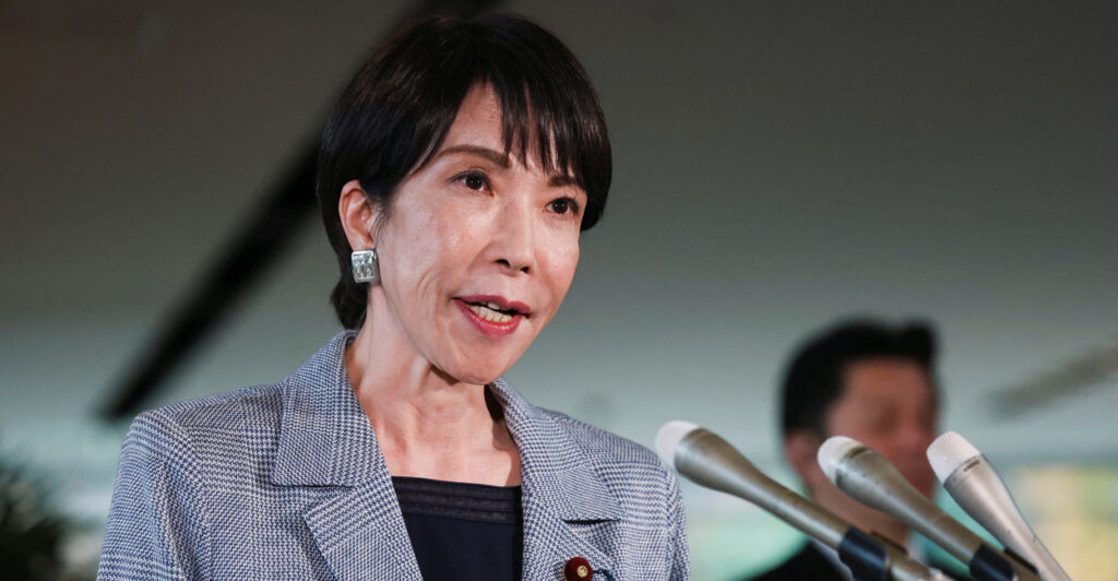 Japanese Prime Minister Sanae Takaichi, in a grey jacket and black shirt, speaks into bank of microphones.