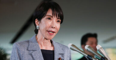 Japanese Prime Minister Sanae Takaichi, in a grey jacket and black shirt, speaks into bank of microphones.