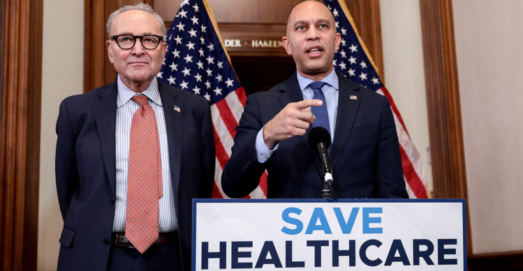 Chuck Schumer stands next to Hakeem Jeffries as he speaks at a podium with the sign "Save Healthcare."