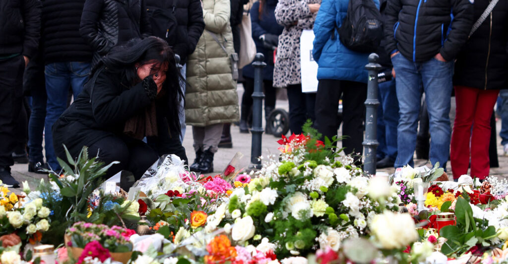 A woman grieves at a flower-strewn memorial as people stand around.