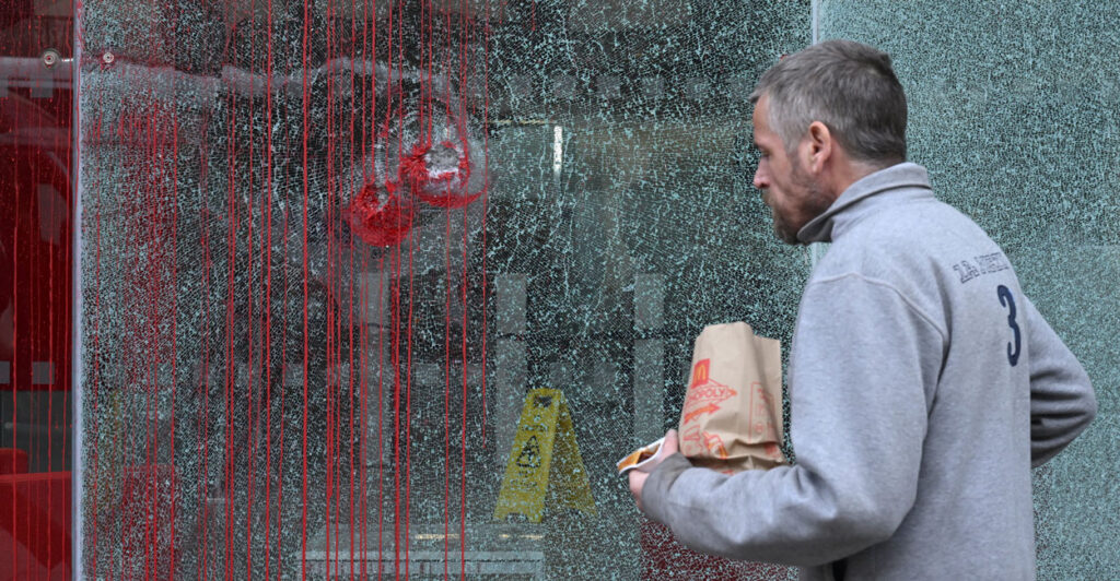 A man in grey jacket walks past a shattered window that's covered in fake blood.