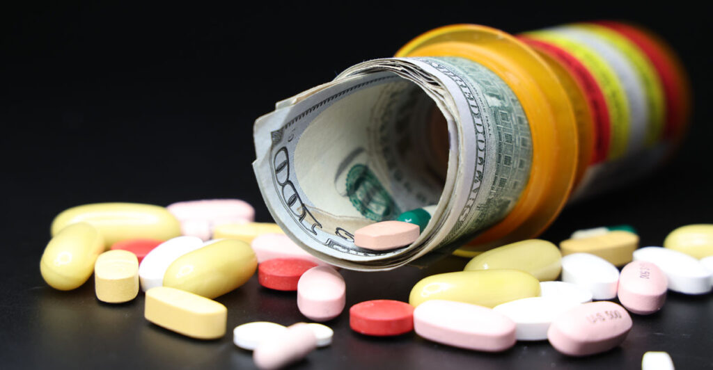 A roll of money sticks out of a pill bottle, with assorted pills laid out before it on a black background.