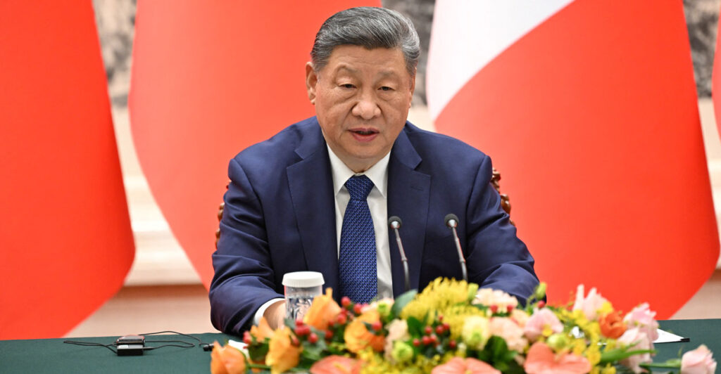Xi Jinping sits at a table in front of a red panel backdrop. In front of him, a sizable flower arrangement.