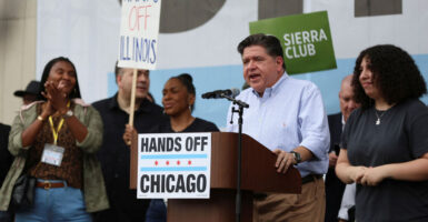 JB Pritzker in a white shirt speaks at a podium that has a sign "Hands Off Chicago."