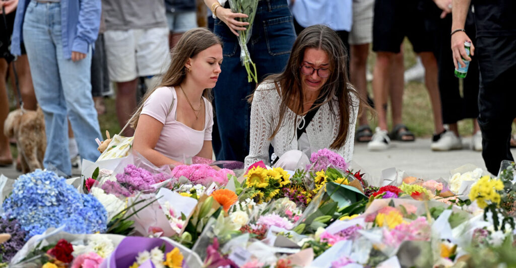 Two women kneel amid a flower memorial.