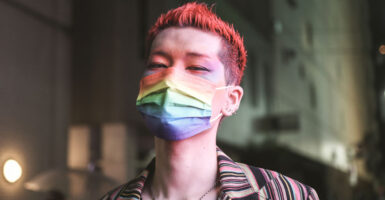 A thin young man with dyed red hair, makeup and a rainbow mask.