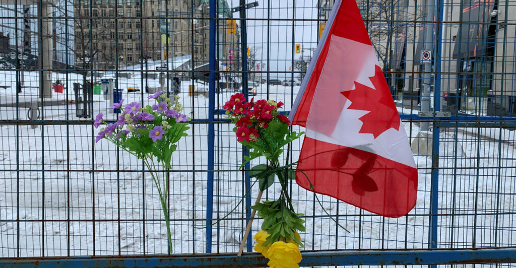 A small Canadian flag and flowers placed against a fence.