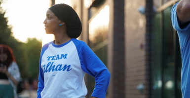 Ihan Omar in a blue-sleeved jersey reading "Team Ilhan" stands on a street.